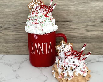 May include: A red ceramic mug with the word "SANTA" in white, topped with faux whipped cream, sprinkles, and a gingerbread snowflake. A smaller decorative item with similar elements sits beside it. Red and white striped straws and the word "Joy" are also included.