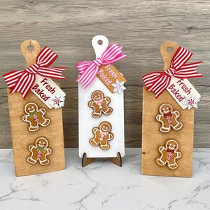 May include: Three decorative wooden cutting boards with gingerbread men. Each board has a bow, a tag that says "Fresh Baked", and snowflake accents. The boards are light brown and white, with red and pink bows.