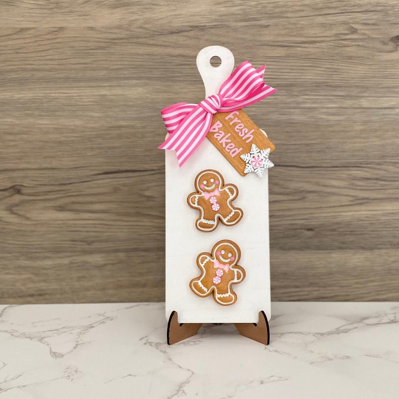 May include: A white wooden cutting board with a pink and white striped bow and two gingerbread man decorations. A wooden tag with the words "Fresh Baked" is attached. The board is supported by a wooden stand.