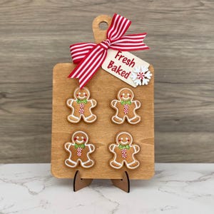 May include: A wooden cutting board with four gingerbread men, a red and white striped bow, and a tag that says "Fresh Baked." The gingerbread men have white icing and green and red accents. The board is on a wooden stand.