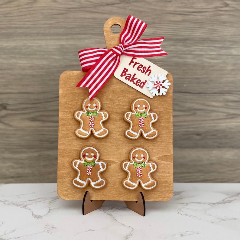 May include: A wooden cutting board with four gingerbread men, a red and white striped bow, and a tag that says "Fresh Baked." The gingerbread men have white icing and green and red accents. The board is on a wooden stand.
