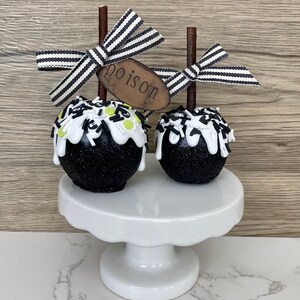Puede incluir: Dos manzanas de caramelo cubiertas de purpurina negra con glaseado blanco y chispas, expuestas sobre un pedestal blanco. Cada manzana tiene un lazo a rayas y una etiqueta de madera que dice "poison". Un dulce de Halloween.