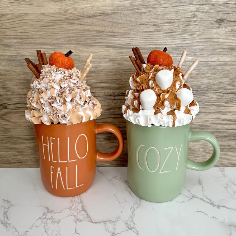 May include: Two decorative mugs with whipped cream toppings, cinnamon sticks, and small pumpkins. One mug is orange with "HELLO FALL" text, the other is green with "COZY" text. The mugs are on a white surface.