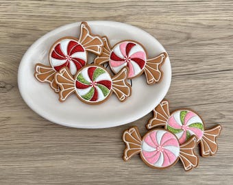 Könnte beinhalten: Auf einem weißen Teller liegen mehrere weihnachtliche Dekorationen in Bonbonform. Die Dekorationen weisen rot, grün und rosa wirbelnde Muster mit braunen Verpackungen auf. Auf einer Holzoberfläche.