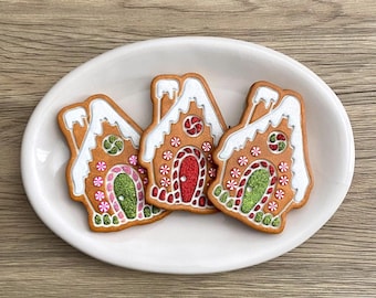 Könnte beinhalten: Drei Lebkuchenhaus-Kekse auf einem weißen, ovalen Teller. Die Kekse haben weiße Zuckergussdächer, rote und grüne Türverzierungen und sind mit bunten Streuseln verziert. Der Teller steht auf einer Holzoberfläche.