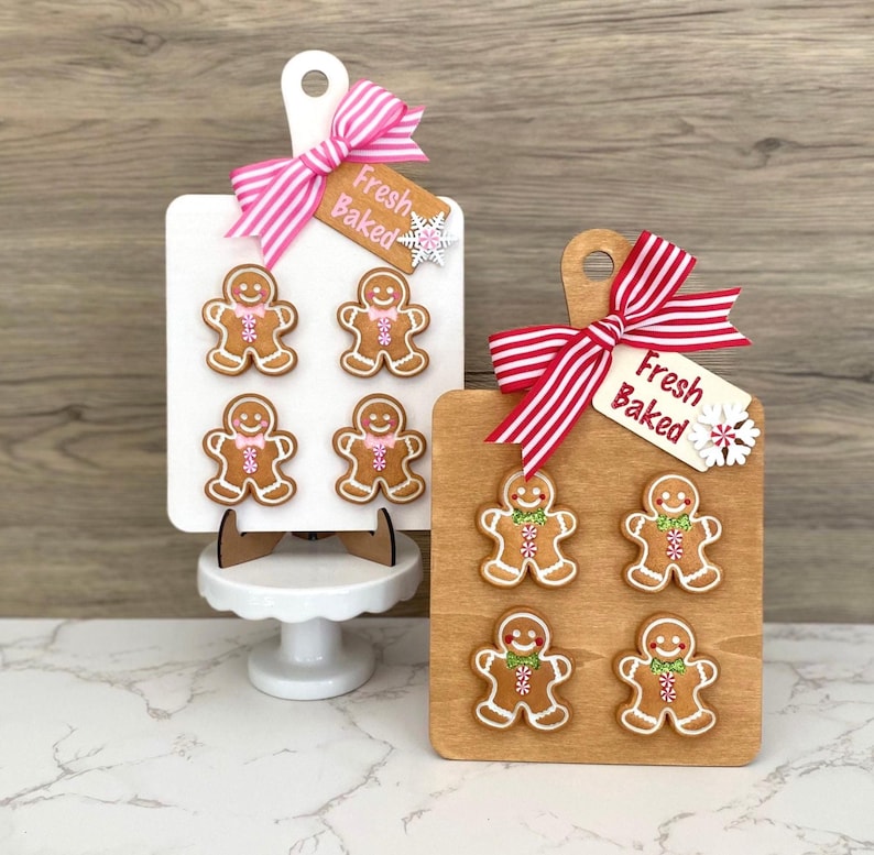 May include: Two decorative wooden cutting boards with gingerbread man designs. One board is white with a pink ribbon, the other is wood-toned with a red and white striped ribbon. Each board has a tag that says "Fresh Baked".