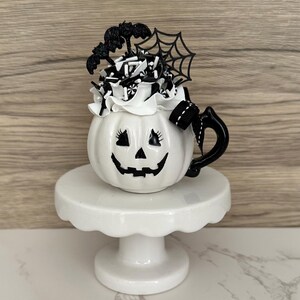 Puede incluir: Una taza blanca de cer&aacute;mica con forma de calabaza con un asa negra, una cara de linterna de Halloween y una parte superior decorativa. La parte superior presenta glaseado blanco, detalles en blanco y negro, un murci&eacute;lago y una telara&ntilde;a. La taza se encuentra sobre un pedestal blanco.