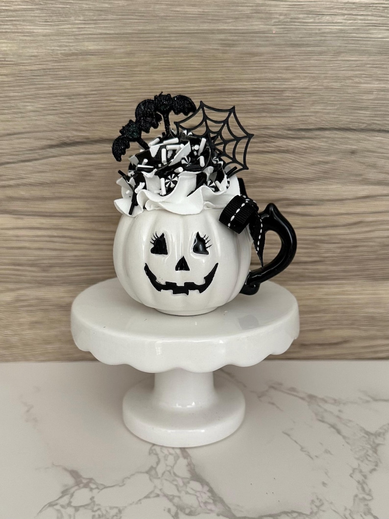Puede incluir: Una taza blanca de cer&aacute;mica con forma de calabaza con un asa negra, una cara de linterna de Halloween y una parte superior decorativa. La parte superior presenta glaseado blanco, detalles en blanco y negro, un murci&eacute;lago y una telara&ntilde;a. La taza se encuentra sobre un pedestal blanco.