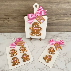 May include: White wooden cutting boards with gingerbread men, pink bows, and tags that read "Fresh Baked." The gingerbread men have pink bow ties and are decorated with white icing. The boards are decorated with snowflake accents.