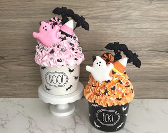 May include: Two Halloween-themed decorative cupcakes. One has pink frosting, a pink ghost, and the word "BOO!" on the cup. The other has orange frosting, a white ghost, and the word "EEK!" on the cup. Both have bat and candy corn accents.