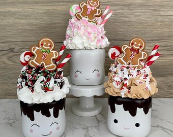 May include: Three white ceramic mugs with smiling faces, decorated to resemble hot chocolate. Each mug features whipped cream, sprinkles, a gingerbread man cookie, and candy canes. The mugs are on a white surface.