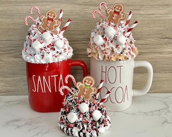 May include: Three decorative mugs with holiday themes. One red mug says "SANTA" and another white mug says "HOT COCOA". Each mug is topped with whipped cream, marshmallows, sprinkles, a gingerbread man, and candy canes. A smaller decorative item is also included.