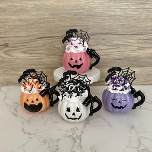Puede incluir: Cuatro tazas de calabaza con tem&aacute;tica de Halloween en rosa, naranja, blanco y morado. Cada taza presenta una cara de jack-o'-lantern, cobertura de crema batida y una decoraci&oacute;n negra de telara&ntilde;a y murci&eacute;lago.