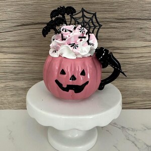 Puede incluir: Una taza de cer&aacute;mica rosa con forma de calabaza con un asa negra y una cara de linterna tallada. La taza est&aacute; llena de crema batida rosa y blanca, decorada con murci&eacute;lagos negros, una telara&ntilde;a y chispas. Se encuentra sobre un pedestal blanco.