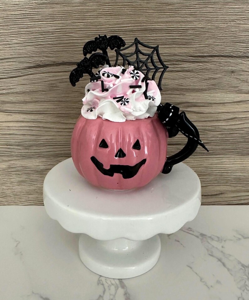 Puede incluir: Una taza de cer&aacute;mica rosa con forma de calabaza con un asa negra y una cara de linterna tallada. La taza est&aacute; llena de crema batida rosa y blanca, decorada con murci&eacute;lagos negros, una telara&ntilde;a y chispas. Se encuentra sobre un pedestal blanco.