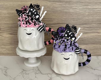May include: Two white ghost-shaped mugs with smiling faces, decorated with pink and purple whipped cream, candy, and black spider accents. One mug sits on a white pedestal, the other has a striped handle.