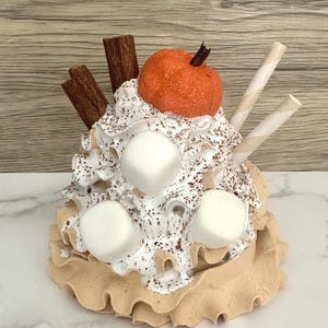 May include: A decorative fall-themed dessert with a pumpkin, marshmallows, cinnamon sticks, and wafer sticks. The dessert has a tan base with a ruffled edge, topped with whipped cream and chocolate sprinkles.