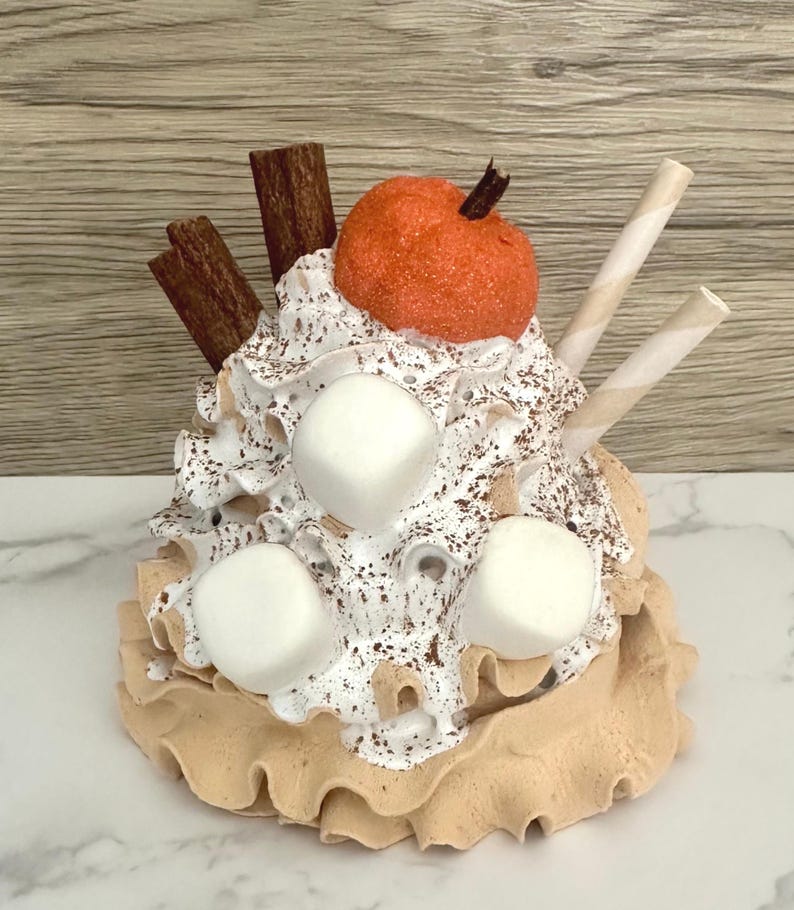 May include: A decorative fall-themed dessert with a pumpkin, marshmallows, cinnamon sticks, and wafer sticks. The dessert has a tan base with a ruffled edge, topped with whipped cream and chocolate sprinkles.
