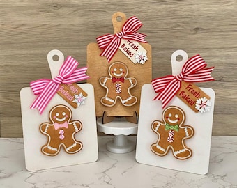 May include: Three decorative cutting boards with gingerbread men. Each board has a ribbon and a tag that says "Fresh Baked". The gingerbread men have different colored bow ties and candy cane accents. The boards are white and light brown.
