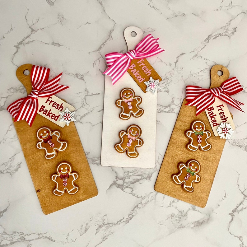 May include: Three wooden cutting boards with gingerbread men and decorative bows. Each board has a gingerbread man design, a tag that says "Fresh Baked", and a decorative bow. The boards are light brown and white.