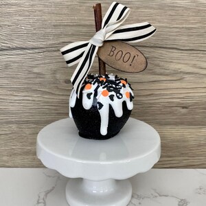 Puede incluir: Una manzana decorativa con tem&aacute;tica de Halloween con una capa de purpurina negra y goteo de glaseado blanco. Est&aacute; rematada con chispas naranjas y negras, un lazo a rayas y una etiqueta que dice "BOO!". La manzana est&aacute; sobre un pedestal blanco.