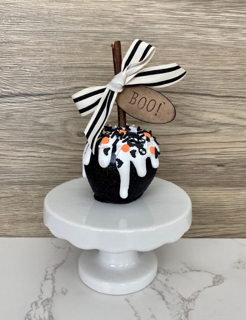 Puede incluir: Una manzana decorativa con tem&aacute;tica de Halloween con una capa de purpurina negra y goteo de glaseado blanco. Est&aacute; rematada con chispas naranjas y negras, un lazo a rayas y una etiqueta que dice "BOO!". La manzana est&aacute; sobre un pedestal blanco.
