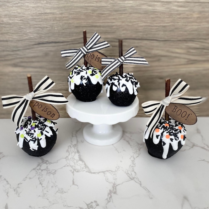 Puede incluir: Manzanas de caramelo negras decoradas para Halloween. Cada manzana tiene un glaseado blanco, chispas y un palito de madera con un lazo a rayas. Algunas manzanas tienen etiquetas de madera que dicen "poison" o "BOO!"