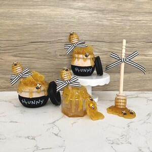 May include: Honey-themed decorative items. Includes honey pots with the word "Hunny", a honey dipper, and faux honey. The honey pots have black and white striped bows and bee accents. The honey appears to be dripping from the pots.