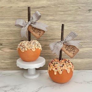 May include: Two decorative caramel apples with a fall theme. Each apple is orange with a caramel drizzle and toppings. They have a brown stick, a gingham bow, and a tag that says "Hello Fall". One apple sits on a white pedestal.