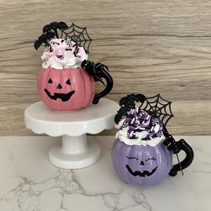 Puede incluir: Dos tazas de cer&aacute;mica con forma de calabaza, una rosa y otra morada, con asas negras y caras de jack-o'-lantern. Cada taza est&aacute; cubierta con crema batida y detalles decorativos, incluyendo murci&eacute;lagos y telara&ntilde;as. La taza rosa est&aacute; sobre un pedestal blanco.