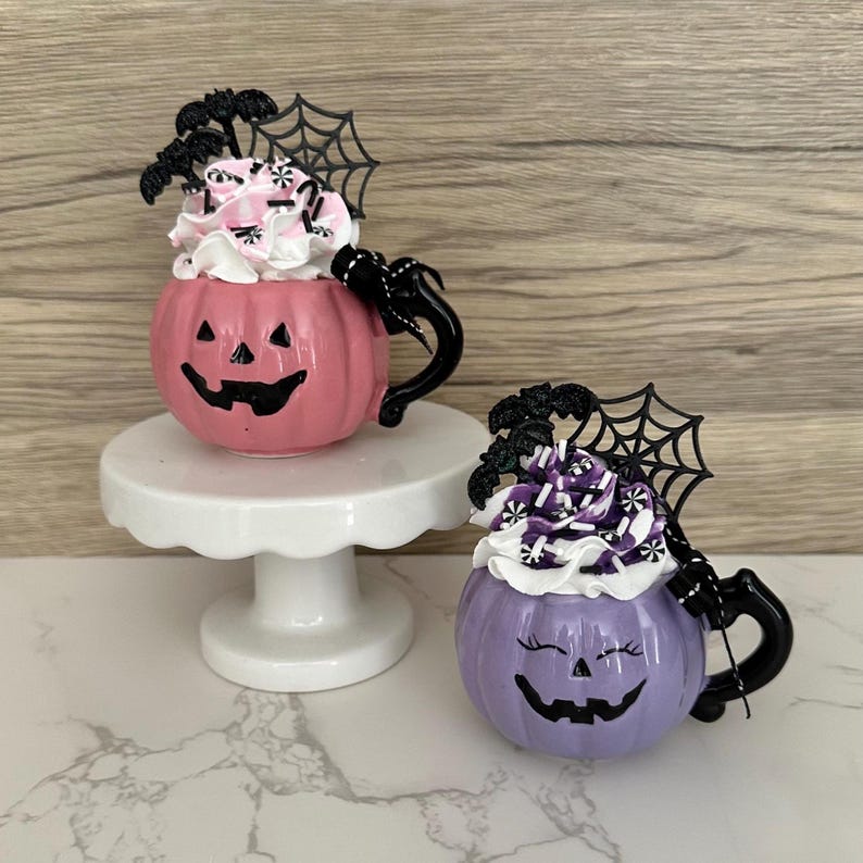 Puede incluir: Dos tazas de cer&aacute;mica con forma de calabaza, una rosa y otra morada, con asas negras y caras de jack-o'-lantern. Cada taza est&aacute; cubierta con crema batida y detalles decorativos, incluyendo murci&eacute;lagos y telara&ntilde;as. La taza rosa est&aacute; sobre un pedestal blanco.
