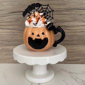 Puede incluir: Una taza de cer&aacute;mica con tem&aacute;tica de Halloween con forma de calabaza con una cara sonriente de jack-o'-lantern. La taza est&aacute; llena de glaseado blanco y naranja, rematada con murci&eacute;lagos decorativos y una telara&ntilde;a. Se encuentra sobre un pedestal blanco.
