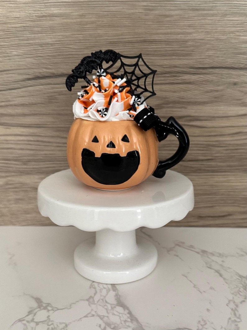 Puede incluir: Una taza de cer&aacute;mica con tem&aacute;tica de Halloween con forma de calabaza con una cara sonriente de jack-o'-lantern. La taza est&aacute; llena de glaseado blanco y naranja, rematada con murci&eacute;lagos decorativos y una telara&ntilde;a. Se encuentra sobre un pedestal blanco.