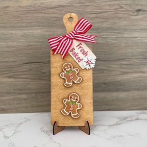 May include: Wooden cutting board with a handle, decorated with two gingerbread men, a red and white striped bow, and a tag that reads "Fresh Baked". The board is propped up on a wooden stand.