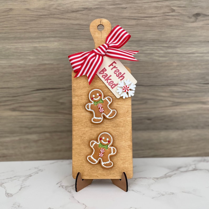May include: Wooden cutting board with a handle, decorated with two gingerbread men, a red and white striped bow, and a tag that reads "Fresh Baked". The board is propped up on a wooden stand.