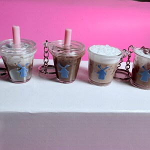 May include: Four miniature coffee drink keychains. Each clear plastic cup features a different coffee drink, including a drink with whipped cream and a drink with chocolate shavings. Each keychain has a pink straw and a blue windmill logo.