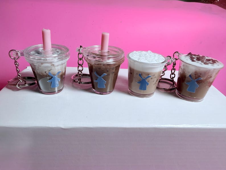 May include: Four miniature coffee drink keychains. Each clear plastic cup features a different coffee drink, including a drink with whipped cream and a drink with chocolate shavings. Each keychain has a pink straw and a blue windmill logo.