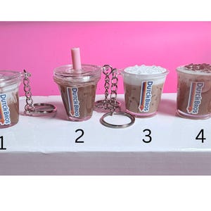 May include: Four miniature Dutch Bros coffee cup keychains. Each clear plastic cup features a different coffee drink, with a pink straw or whipped cream topping. The Dutch Bros logo is visible on each cup. The keychains are attached with silver chains and rings.