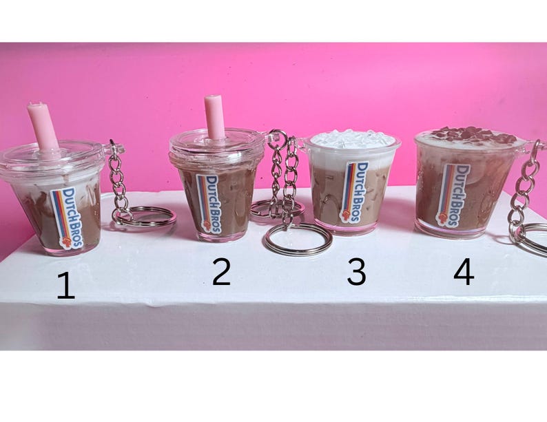 May include: Four miniature Dutch Bros coffee cup keychains. Each clear plastic cup features a different coffee drink, with a pink straw or whipped cream topping. The Dutch Bros logo is visible on each cup. The keychains are attached with silver chains and rings.