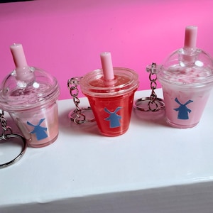 May include: Three clear plastic drink cup keychains with pink liquid and a blue windmill logo. Each keychain has a pink straw and a silver chain with a keyring. The background is pink and the keychains are on a white surface.
