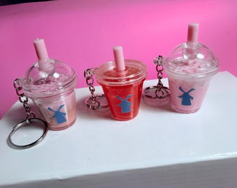 May include: Three clear plastic drink cup keychains with pink liquid and a blue windmill logo. Each keychain has a pink straw and a silver chain with a keyring. The background is pink and the keychains are on a white surface.