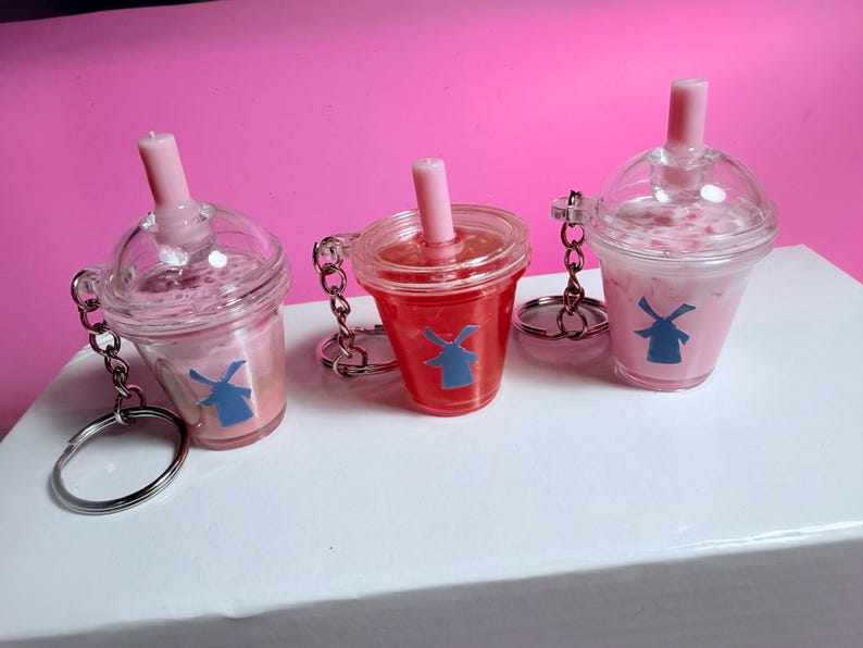 May include: Three clear plastic drink cup keychains with pink liquid and a blue windmill logo. Each keychain has a pink straw and a silver chain with a keyring. The background is pink and the keychains are on a white surface.