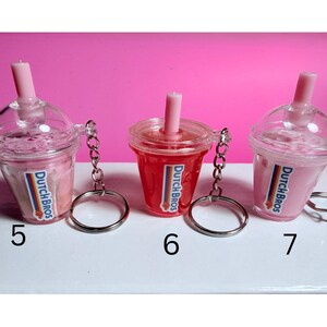 May include: Three miniature Dutch Bros drink keychains. Two are pink, one is red, all with a pink straw and the Dutch Bros logo. Each keychain has a silver ring attached. Numbers 5, 6, and 7 are visible.