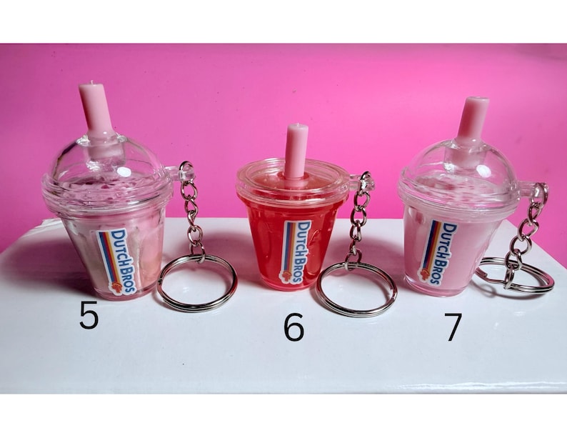 May include: Three miniature Dutch Bros drink keychains. Two are pink, one is red, all with a pink straw and the Dutch Bros logo. Each keychain has a silver ring attached. Numbers 5, 6, and 7 are visible.