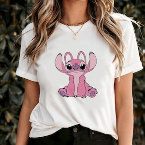 May include: White t-shirt featuring a pink cartoon character with large ears and a friendly expression. The character is sitting, with its arms raised. The shirt has short sleeves and a crew neck.