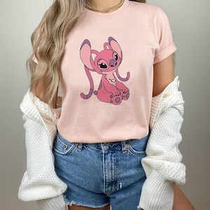 May include: A light pink t-shirt featuring a cartoon character in shades of pink and purple. The character has large ears and is sitting down. The shirt is paired with denim shorts and a white cardigan.
