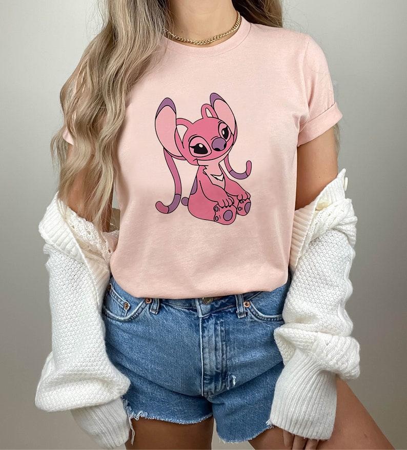 May include: A light pink t-shirt featuring a cartoon character in shades of pink and purple. The character has large ears and is sitting down. The shirt is paired with denim shorts and a white cardigan.