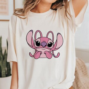 May include: Cream-colored t-shirt featuring a pink cartoon character with large ears and black eyes. The character is centered on the shirt, with a simple, clean design. The shirt has a relaxed fit.