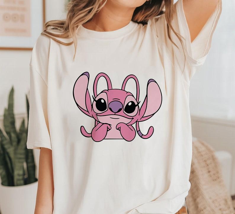 May include: Cream-colored t-shirt featuring a pink cartoon character with large ears and black eyes. The character is centered on the shirt, with a simple, clean design. The shirt has a relaxed fit.