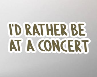 May include: A white sticker with the text "I'D RATHER BE AT A CONCERT" in a brown, handwritten-style font. The text is outlined in white, creating a layered effect. The sticker is likely for personalizing items.